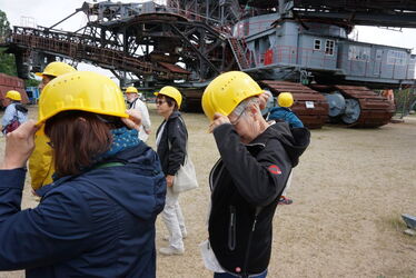 Exkursionsteilnehmer setzen Helme auf. Im Hintergrund sieht man den Großraumbagger Hagenwerder